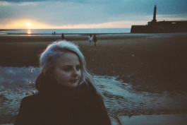 ID: A grainy film photograph of Lizzy with long bleached hair in profile on Margate beach. In the background we see wet sand and the sea in the distance with the end of the pier to the right of the shot.
