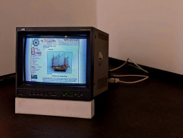 Photograph of a broadcast monitor resting on a white prop, which raises it off the florr at the front.On screen is an screenshot taken from a website from the late 2000s, showing a sales listing for a model sailing boat. 