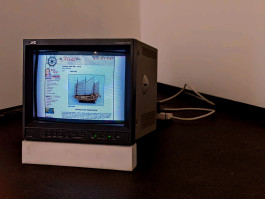 Photograph of a broadcast monitor resting on a white prop, which raises it off the florr at the front.On screen is an screenshot taken from a website from the late 2000s, showing a sales listing for a model sailing boat.