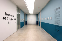 A photograph of a long, tall and wide corridor. The right hand wall and half of the left wall are painted in two tones of blue, similar to those found in hospitals. There are small artworks in white frames on the wall, but we can't see the detail from this vantage point. The nearest part of the left hand wall is white and has written on it, in large, cursive text the following: Remember, you are sick, but that's okay. 