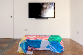 Photograph showing Sick Blue Sea as shown at Crate in 'Things I Have Learned the Hard Way'. This view shows a large flatscreen monitor and a bench positioned for viewing the film on the monitor, which is covered with red, white, blue and green plastic bags. 