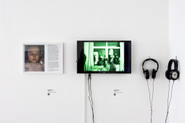 Photograph showing two artworks installed in a white-walled gallery. On the left is a photograph in a white frame - 'Bathtub Selfie', details of which can be seen below. On the right, a flatscreen monitor with two sets of headphones attached to the wall. The screen is showing a still from 'Feeling The Fantasy', details below. 